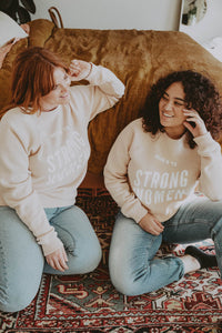 Two women sat on the floor by a bed wearing Here's To Strong Women Sweatshirt in Unisex by The Bee and The Fox