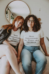 Two women sat on a bed wearing Never Underestimate the Power of a Woman Shirt in white by The Bee and The Fox