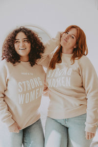 Two women wear Here's To Strong Women Sweatshirt in Unisex by The Bee and The Fox